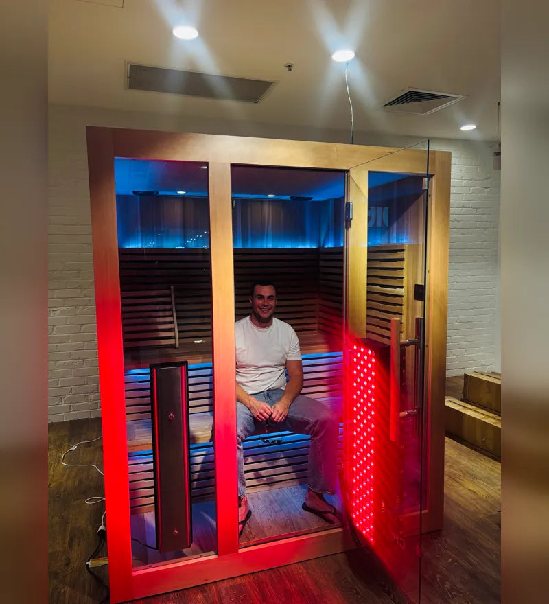 Person sitting in an infrared sauna with a red light therapy panel glowing beside them