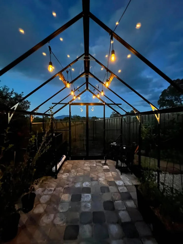 Inside a customer's Wintergardenz 8ft Wide Glasshouse at dusk, strung with festoon lights over a tiled floor